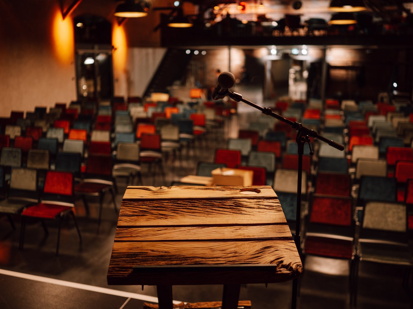 Vordergrund ein Holzpult vor einer Reihenbestuhlung mit bunten Stühlen
